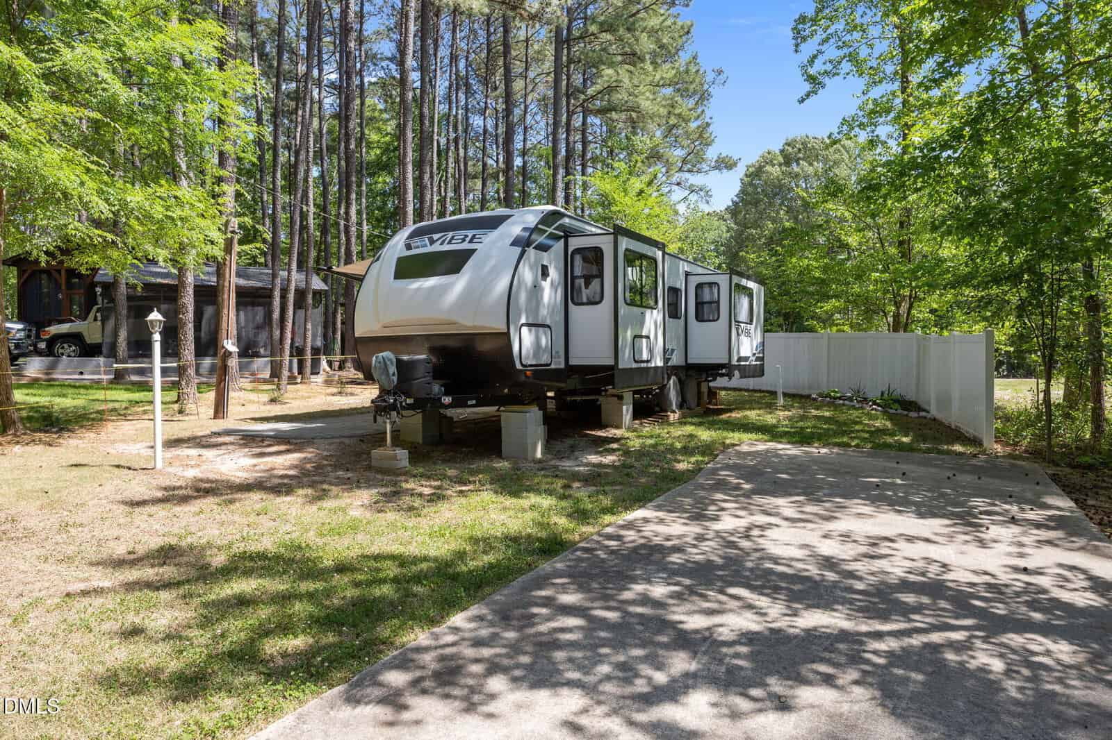 A white Vibe travel trailer is parked on a grassy lot surrounded by tall trees, next to a concrete driveway and a white fence, with sunlight filtering through the leaves.