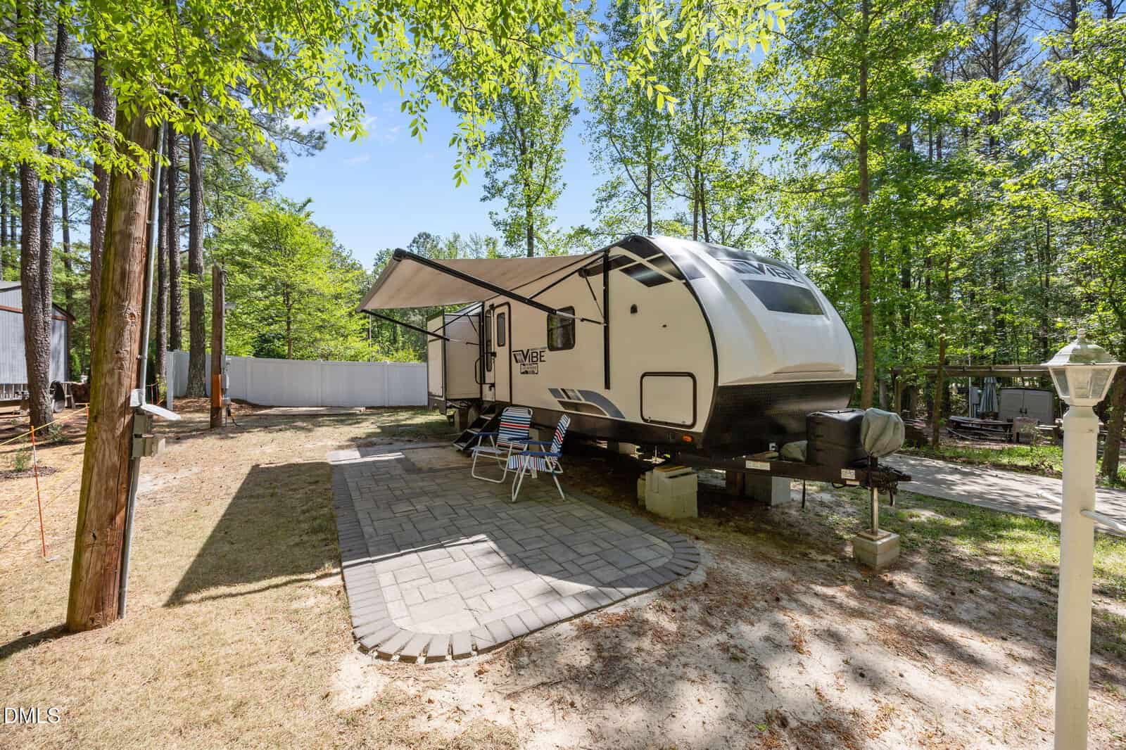 A travel trailer is parked on a paved pad in a wooded area with its awning extended. Two lawn chairs and a small table sit under the awning, surrounded by tall trees and dappled sunlight.