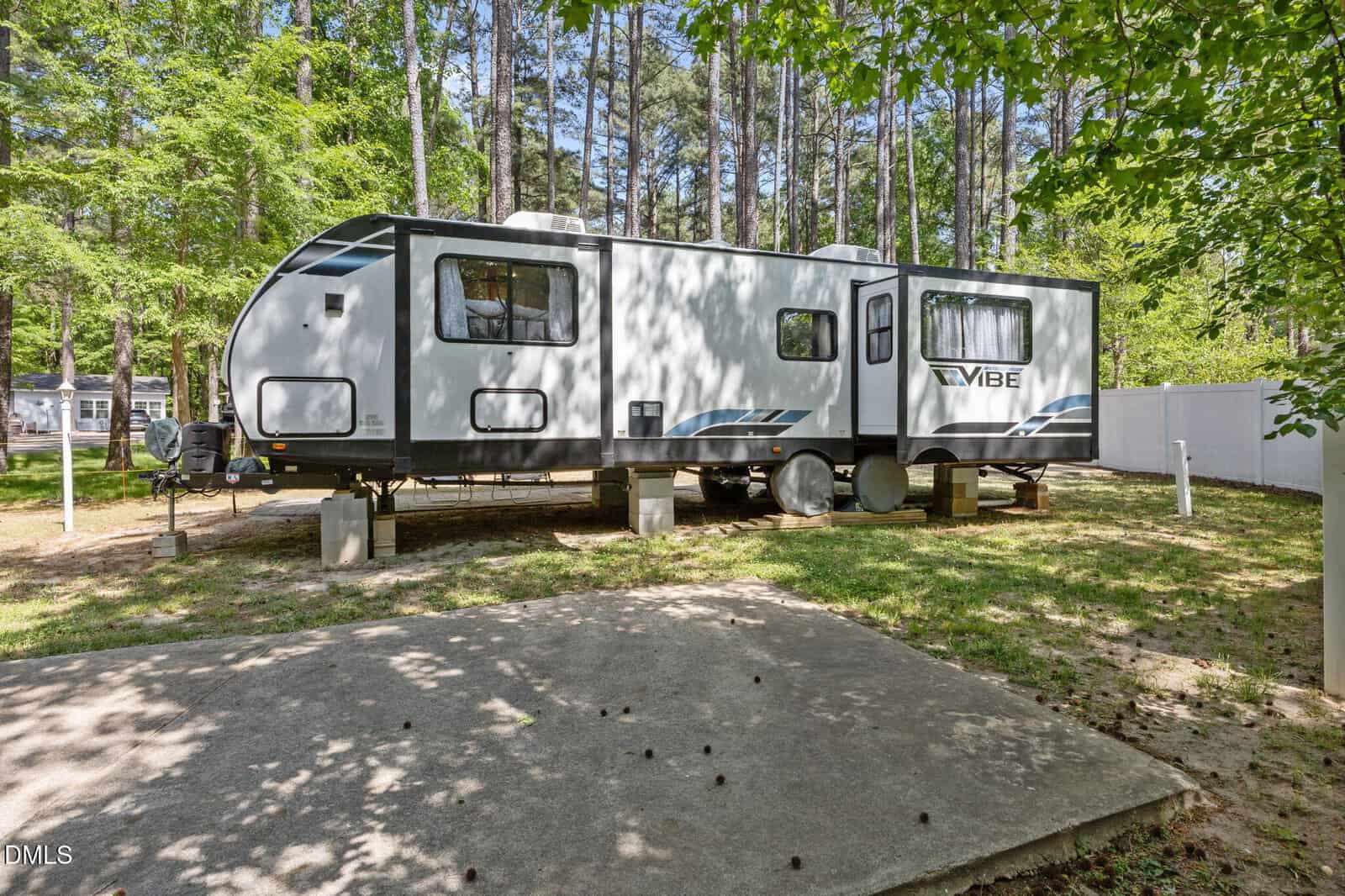 A large white and gray travel trailer is parked on concrete blocks in a wooded area with tall trees and a white fence in the background. Sunlight filters through the leaves, casting shadows on the ground.