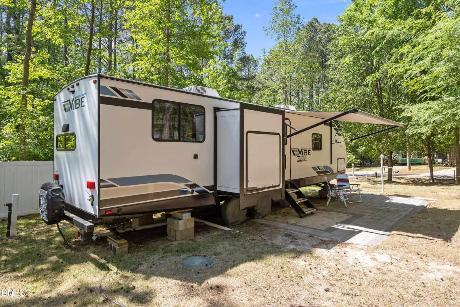 A white and gray RV with an extended awning is parked on a shaded lot surrounded by trees. Patio furniture, including blue chairs and a small table, sits on the concrete area beside the RV.