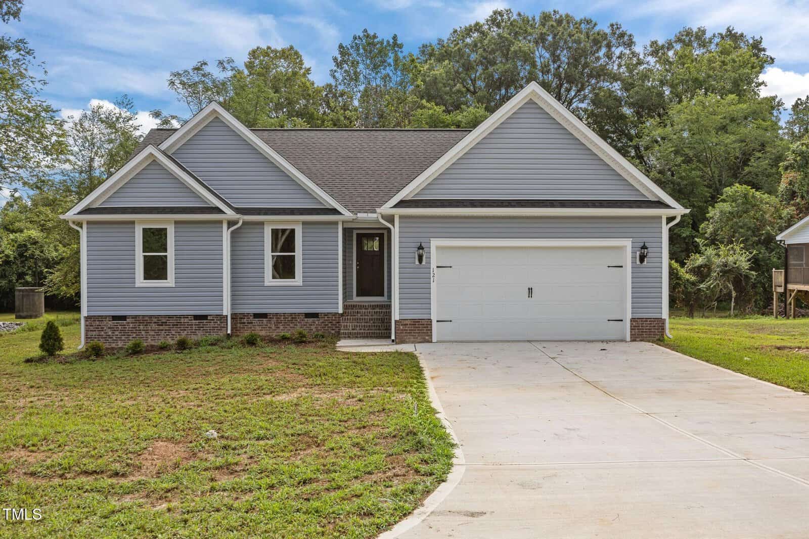 Single-story house with light gray siding, white trim, dark roof shingles, a brick accent near the foundation, and a two-car garage. The front yard has sparse grass and a wide concrete driveway. Trees are in the background.
