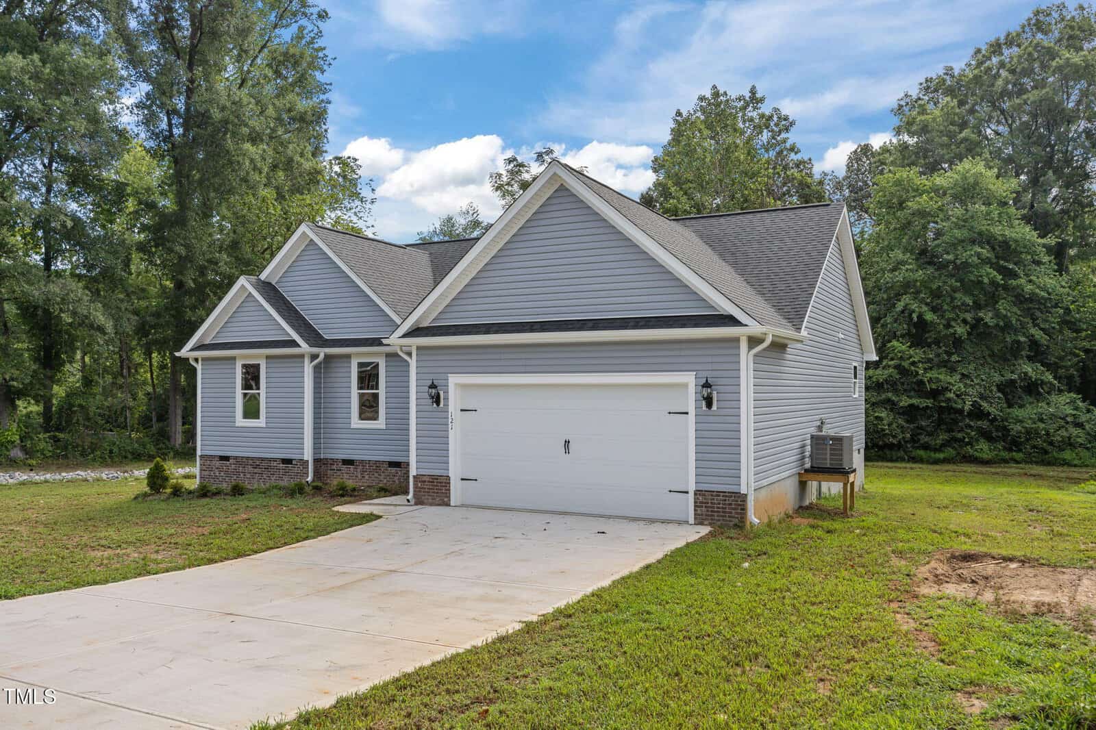 A single-story, gray house with a gabled roof, white trim, and a double garage sits on a concrete driveway, surrounded by green grass and trees under a partly cloudy sky.