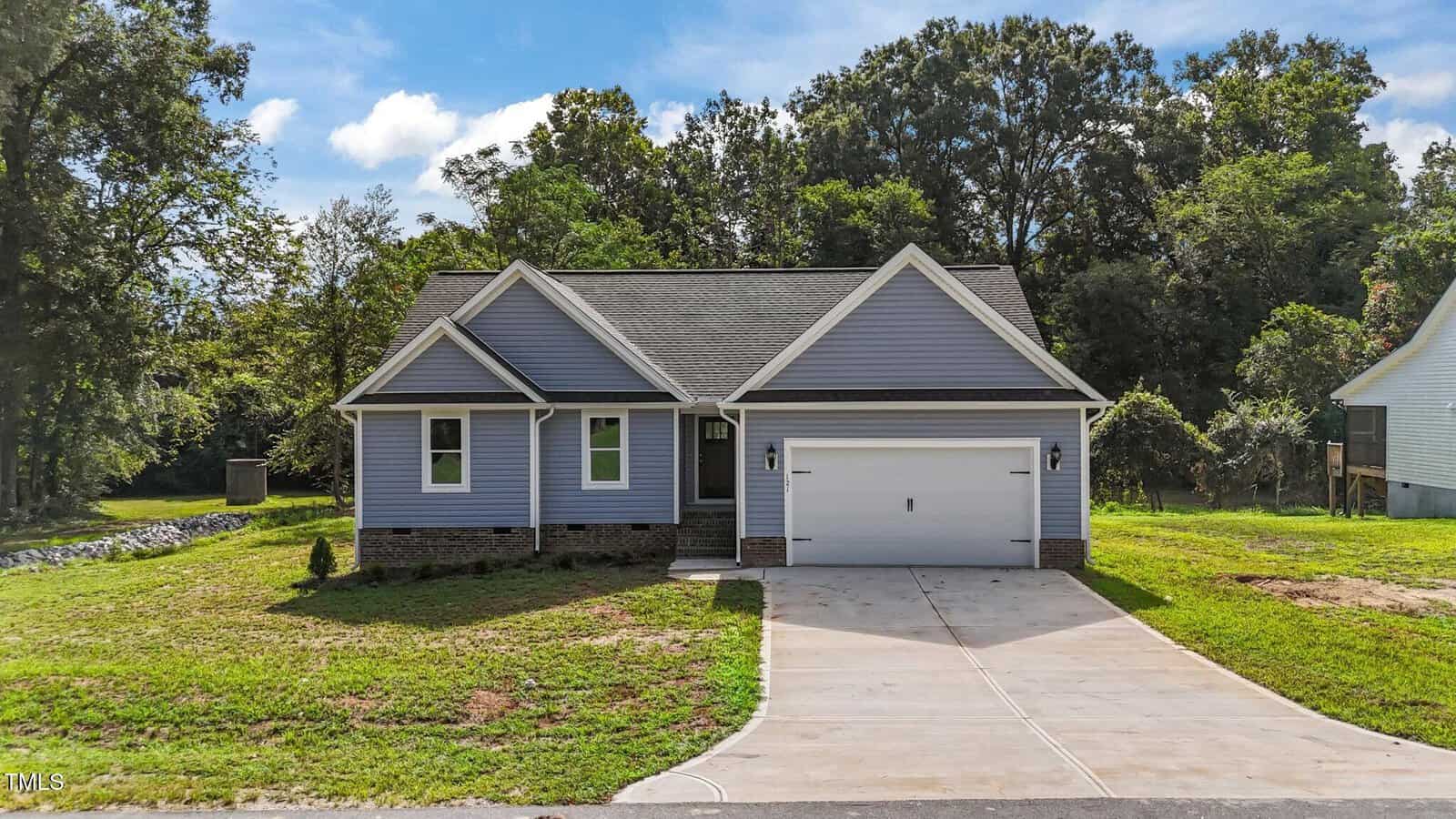 Single-story house with gray siding, a gabled roof, attached two-car garage, and a concrete driveway. The home is surrounded by a green lawn and trees under a partly cloudy sky.