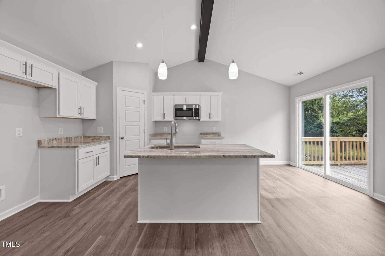 Modern kitchen with white cabinets, an island with a sink, stainless steel appliances, pendant lights, wood flooring, and sliding glass doors leading to a deck with a view of trees outside.