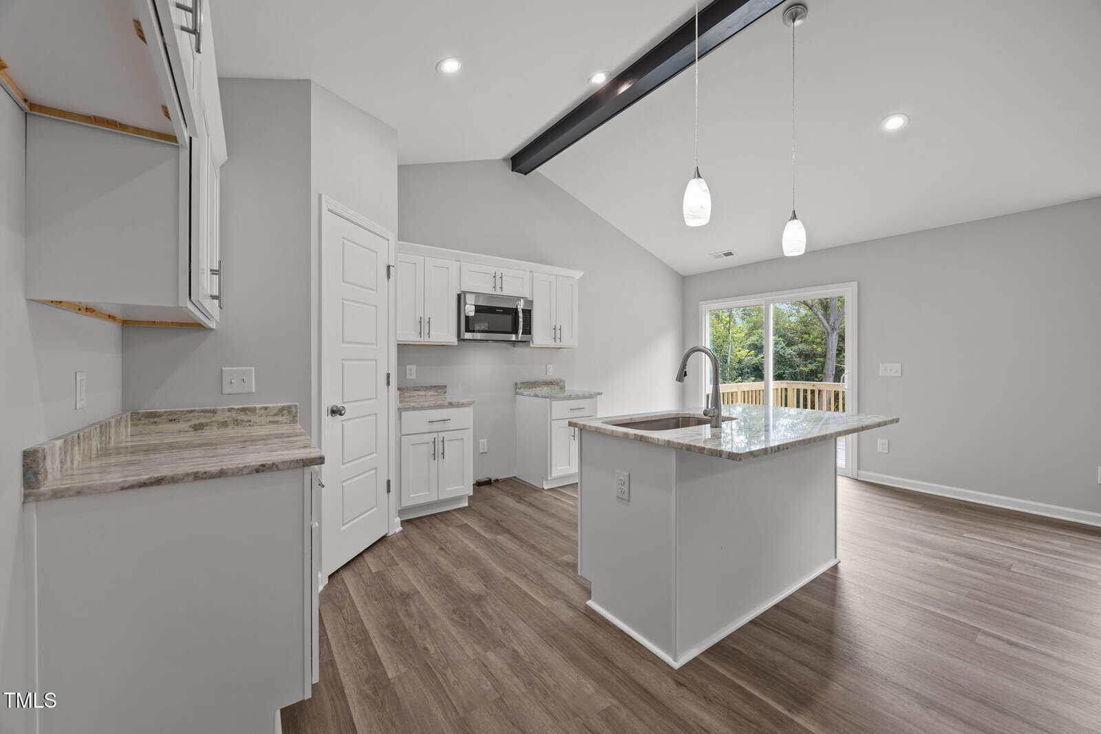 Modern kitchen with wood floors, white cabinets, stainless steel appliances, island with sink, pendant lights, and sliding glass doors leading to a deck with a view of green trees.