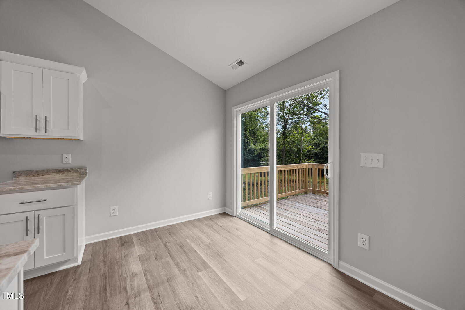 A bright, empty kitchen area with light wood flooring, white cabinets, and sliding glass doors opening to a wooden deck with a view of green trees outside.