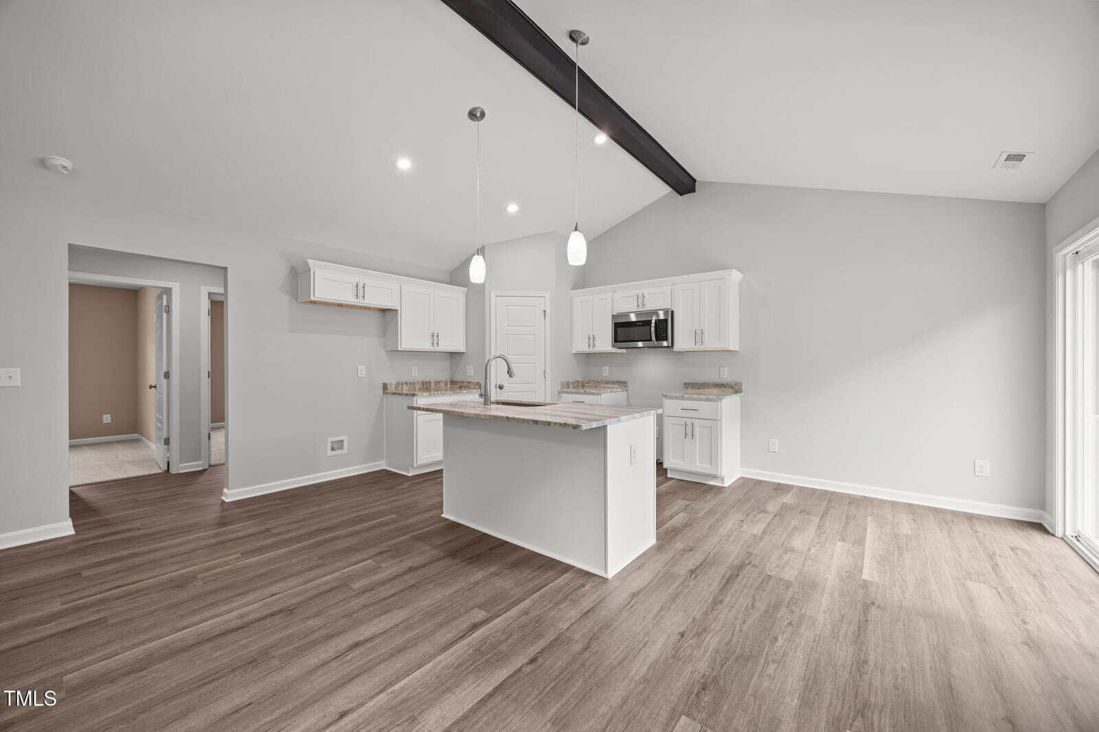 Modern kitchen with white cabinets, a center island, pendant lights, stainless steel appliances, wood floors, vaulted ceiling with dark beam, and neutral walls. A doorway leads to other rooms and a sliding door brings in natural light.