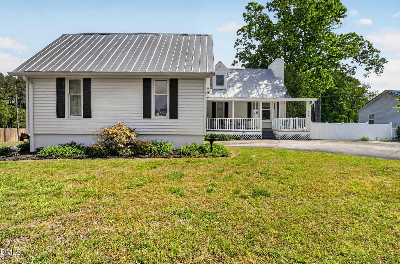 A white house with black shutters, a metal roof, covered front porch, and well-kept lawn on a sunny day. A concrete driveway curves in front of the house, and trees and shrubs border the yard.