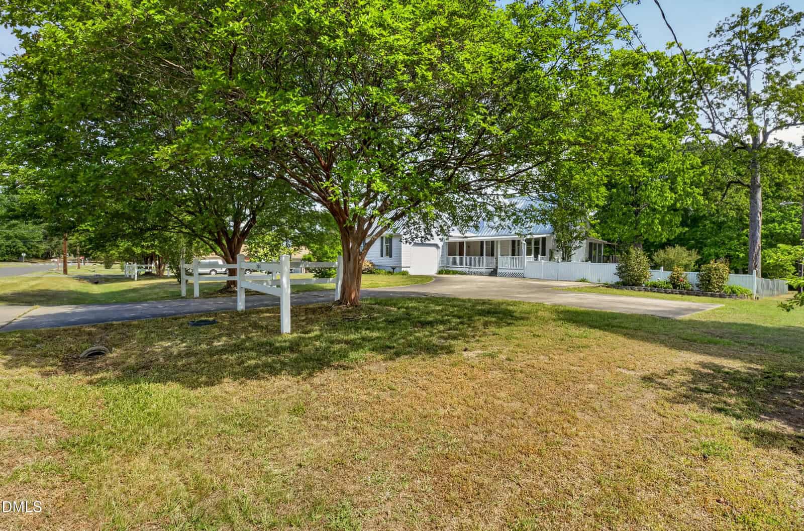 A white house with a large driveway is partially hidden behind green trees and a white fence, with grass and a street in the foreground on a sunny day.