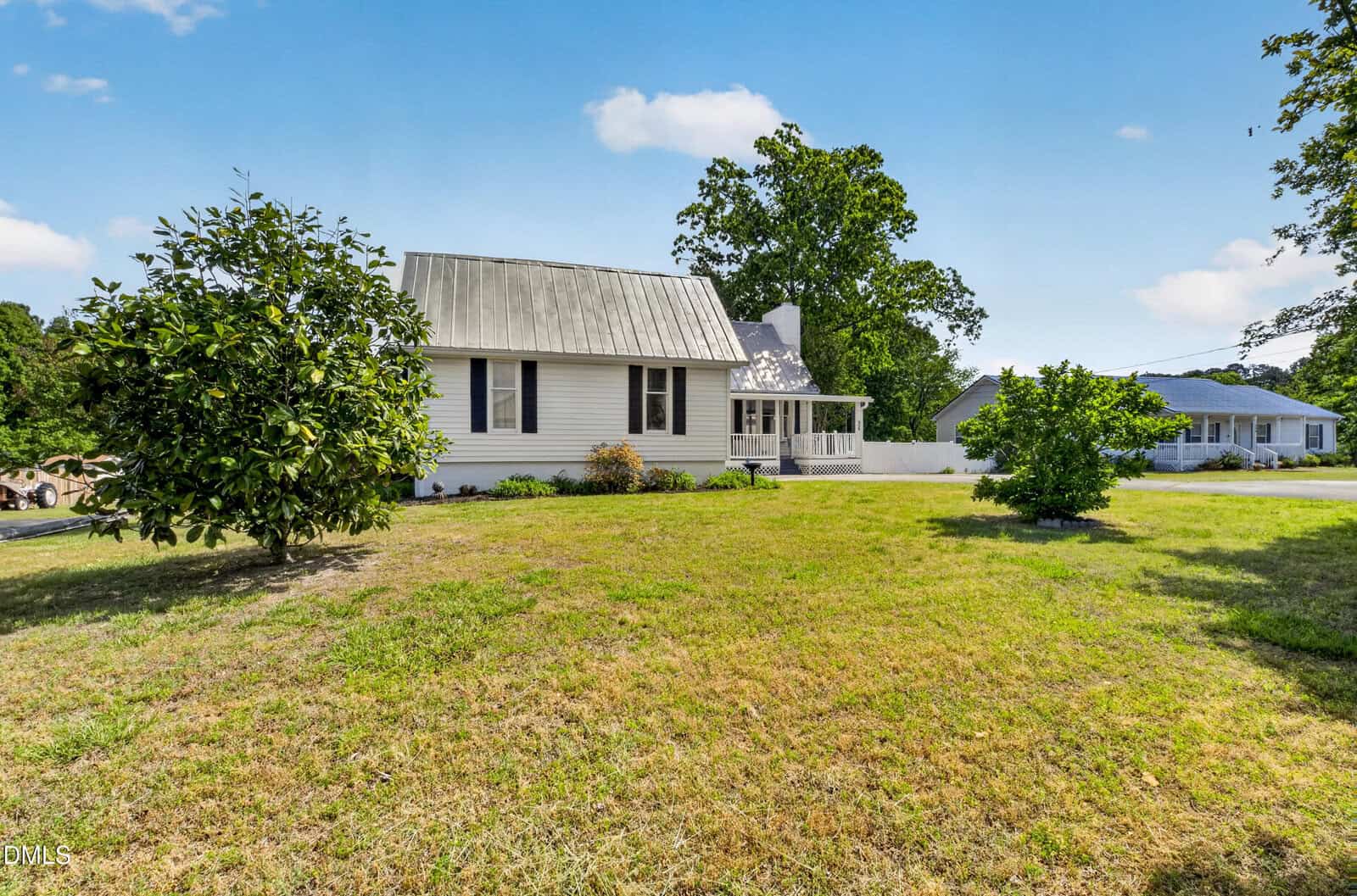 White house with a metal roof and front porch sits on a large, grassy yard with two small trees. Another house is visible in the background, under a bright blue sky with a few clouds.