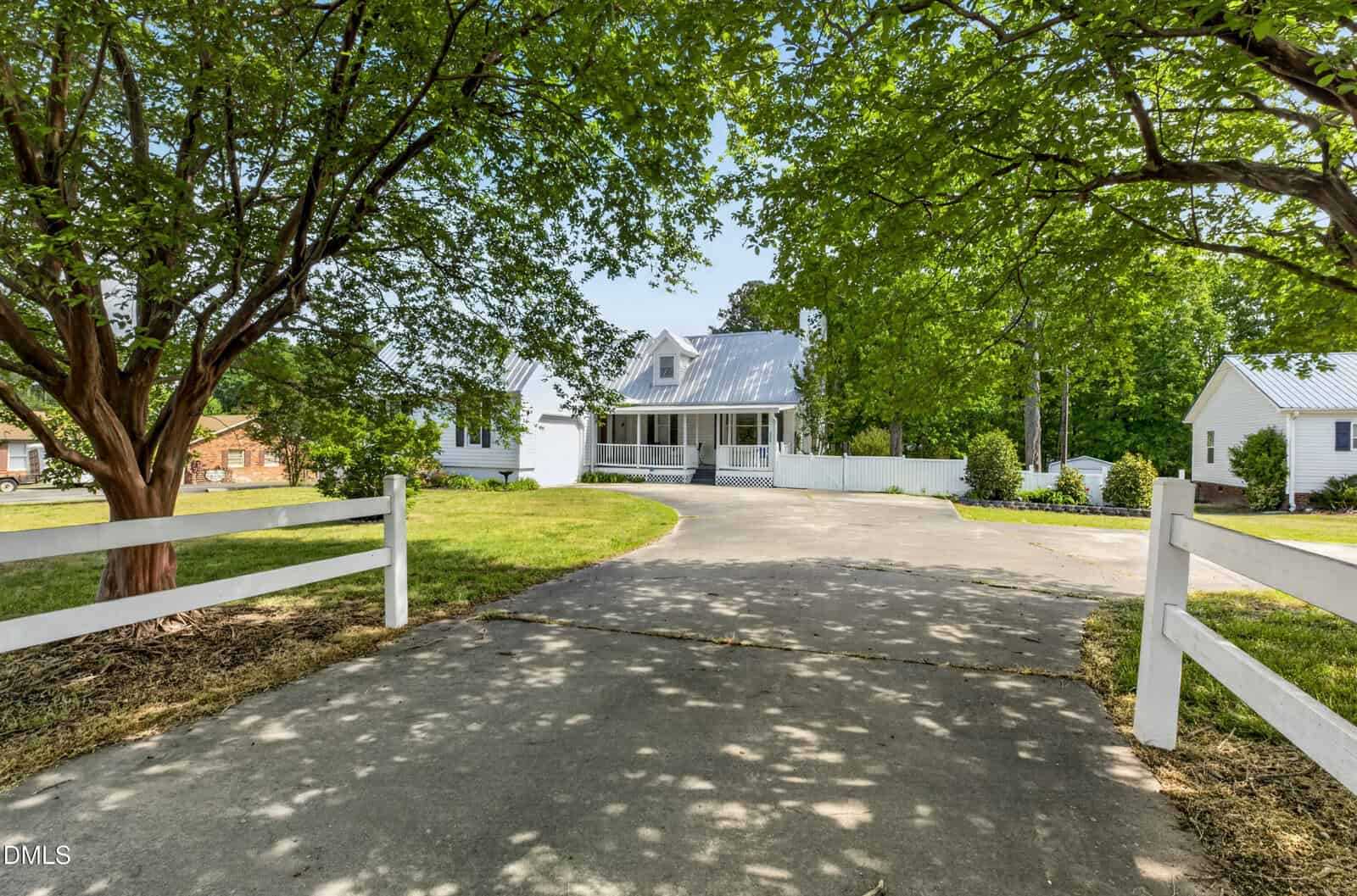 A white house with a front porch and metal roof sits at the end of a curving driveway, surrounded by green trees and a white fence under a clear sky.