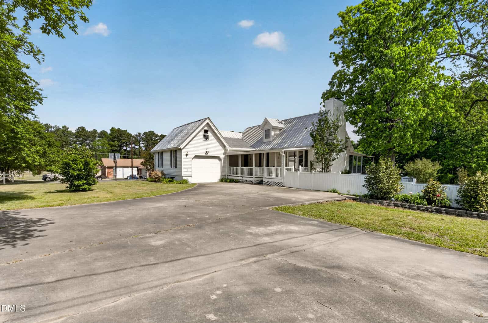 A white house with a metal roof, attached garage, large front porch, and a wide driveway, surrounded by green trees and grass under a blue sky.