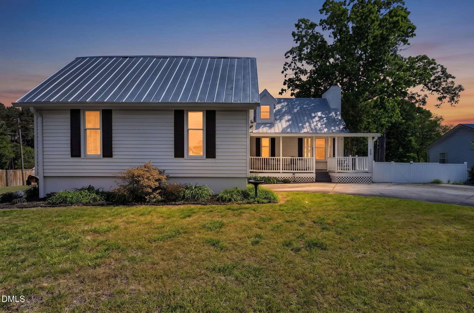 A charming white house with black shutters and a metal roof at sunset, featuring a covered front porch, a well-kept lawn, and a tree in the background. Warm lights glow from the windows.