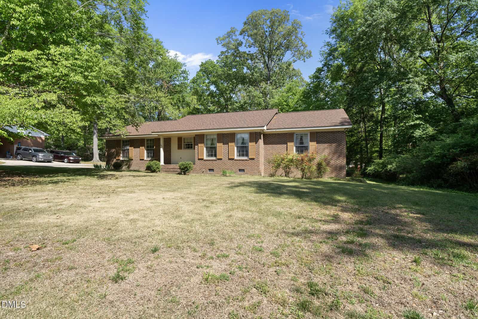 A single-story brick house with white-trimmed windows and a brown roof sits on a spacious lawn, surrounded by green trees under a clear blue sky.