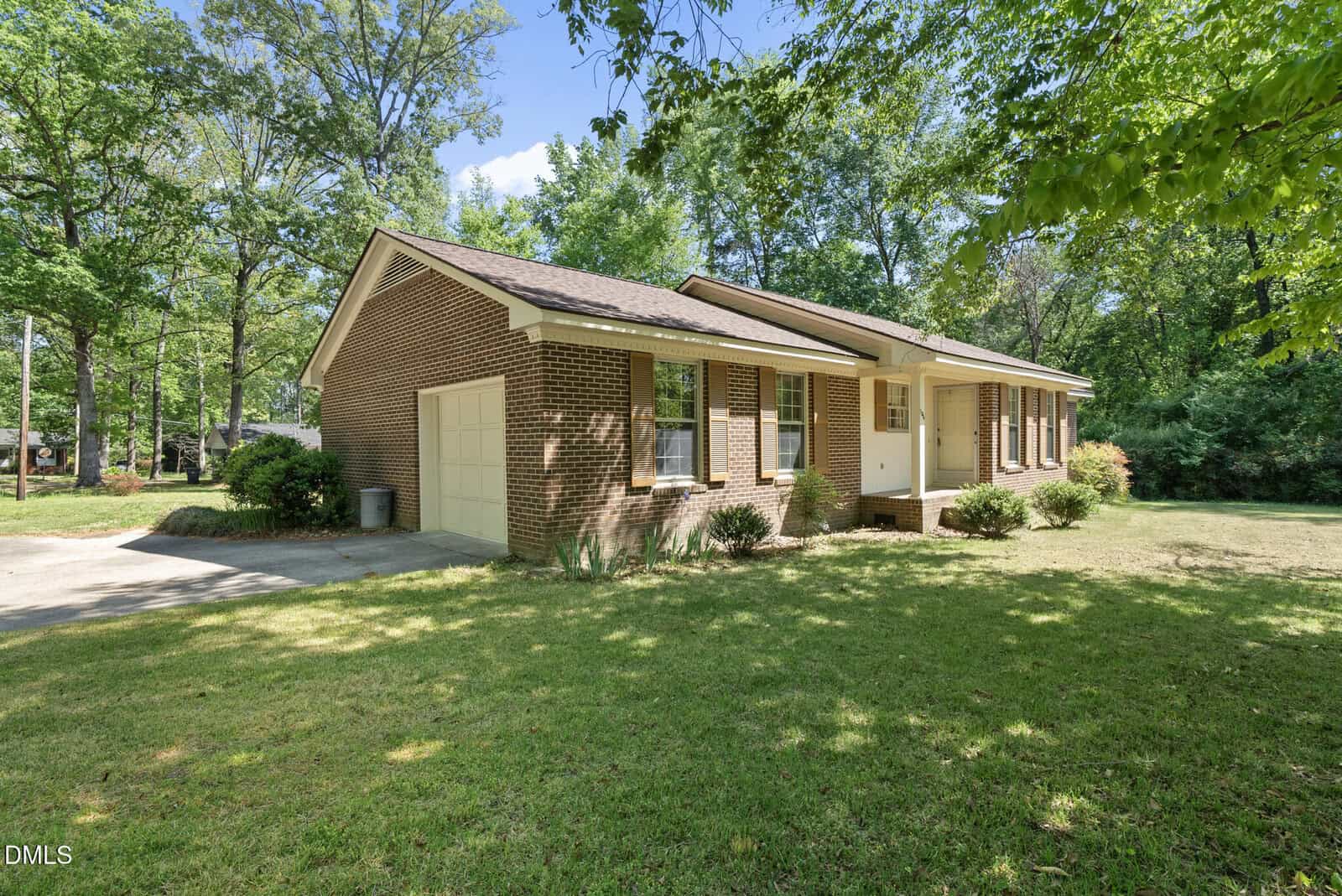 A single-story brick house with an attached garage, surrounded by green grass, bushes, and tall trees under a blue sky on a sunny day.