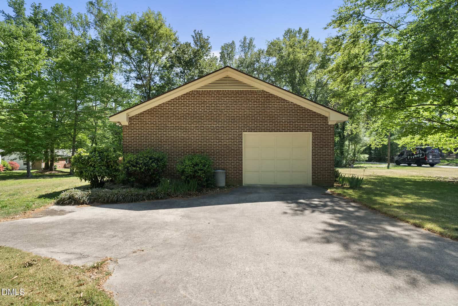 A single-car garage with a brown brick exterior, a light-colored garage door, and a concrete driveway, surrounded by green trees and shrubs on a sunny day.