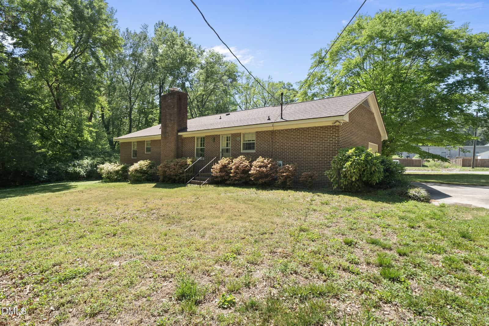 A single-story brick house with a chimney sits on a grassy lot surrounded by trees. There are shrubs along the front and steps leading to the entrance. The sky is clear and sunny.