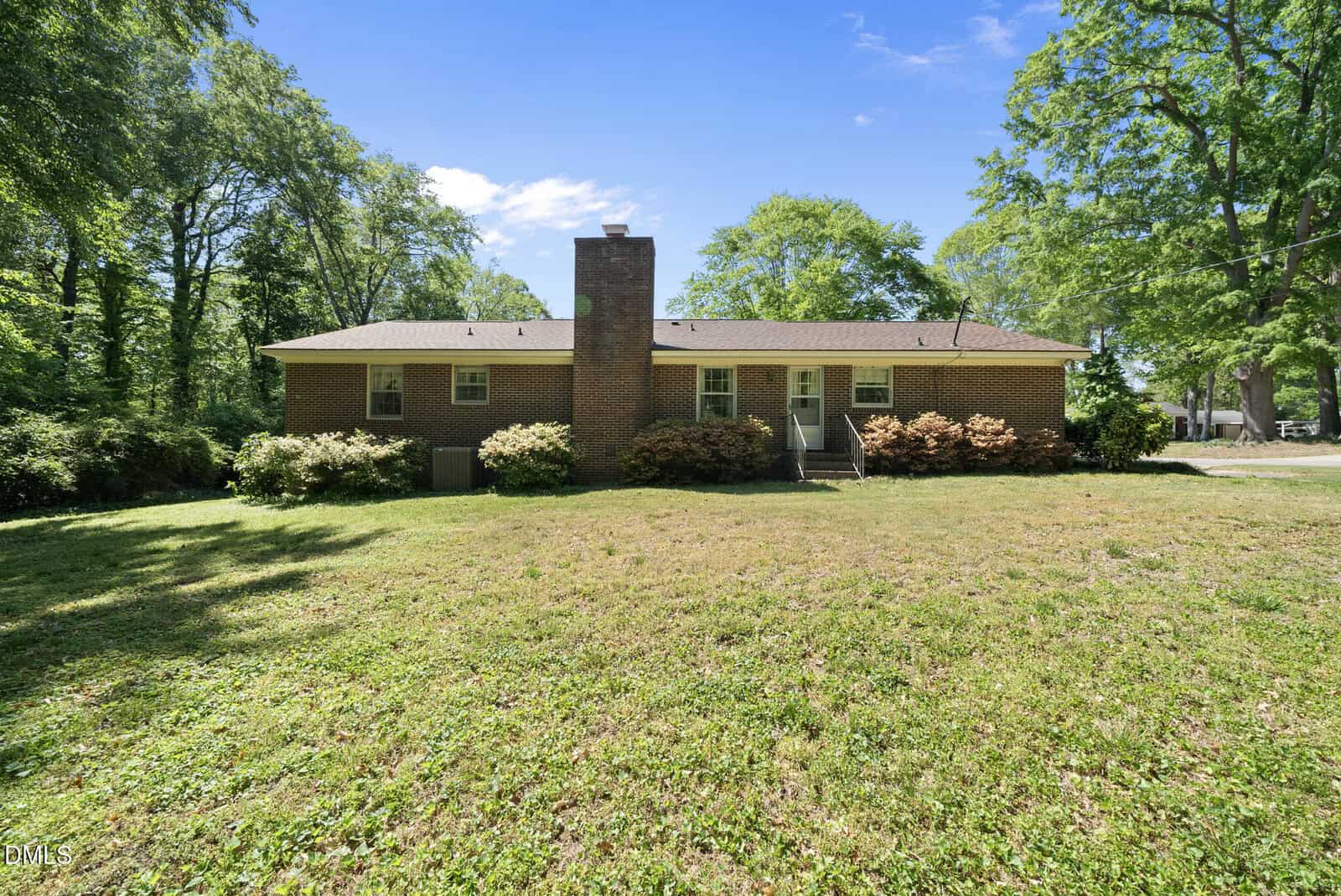 A single-story brick house with a central chimney, surrounded by bushes and trees, sits behind a large, grassy front yard under a clear blue sky.