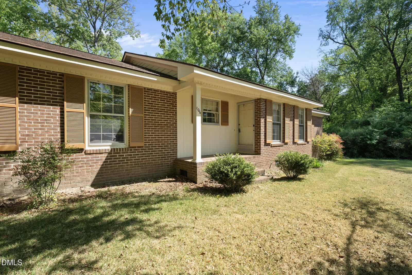 Single-story brick house with tan shutters, white trim, small covered porch, and shrubs in front. The yard has dry grass and is surrounded by tall green trees under a blue sky.