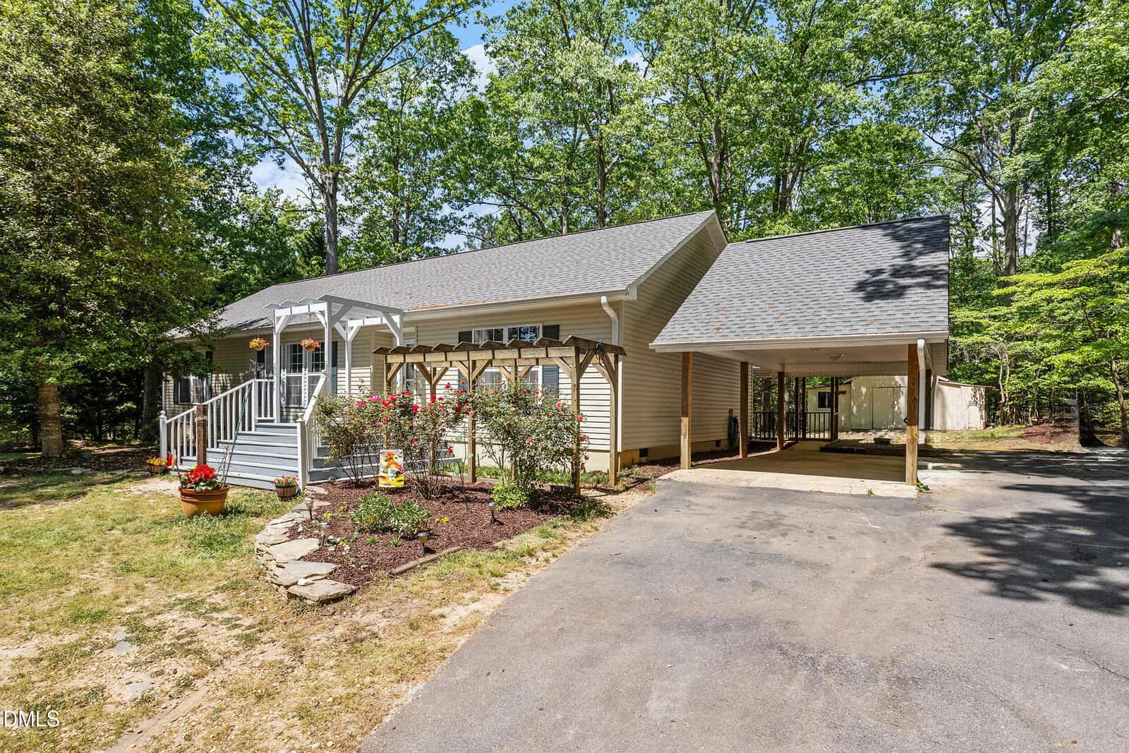 Single-story beige house with a front porch, white railings, landscaped flower beds, and a covered carport. Surrounded by tall green trees and a paved driveway on a sunny day.