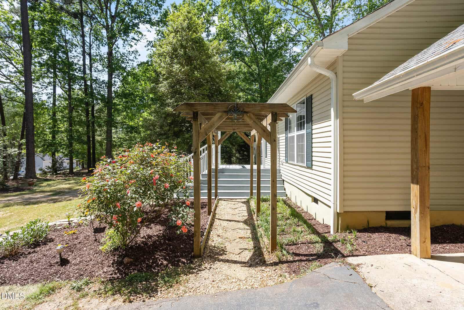 A wooden arbor stands over a gravel path beside a tan house with white trim, leading to a deck. Blooming rose bushes and mulch line the path, and tall trees surround the yard on a sunny day.