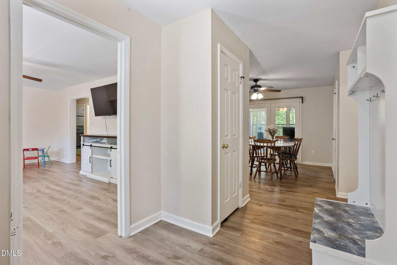 View of a hallway leading to a dining area with a round table and chairs. The living room is visible to the left, featuring a wall-mounted TV and a children’s chair. Walls are light-colored, and floors are wood.