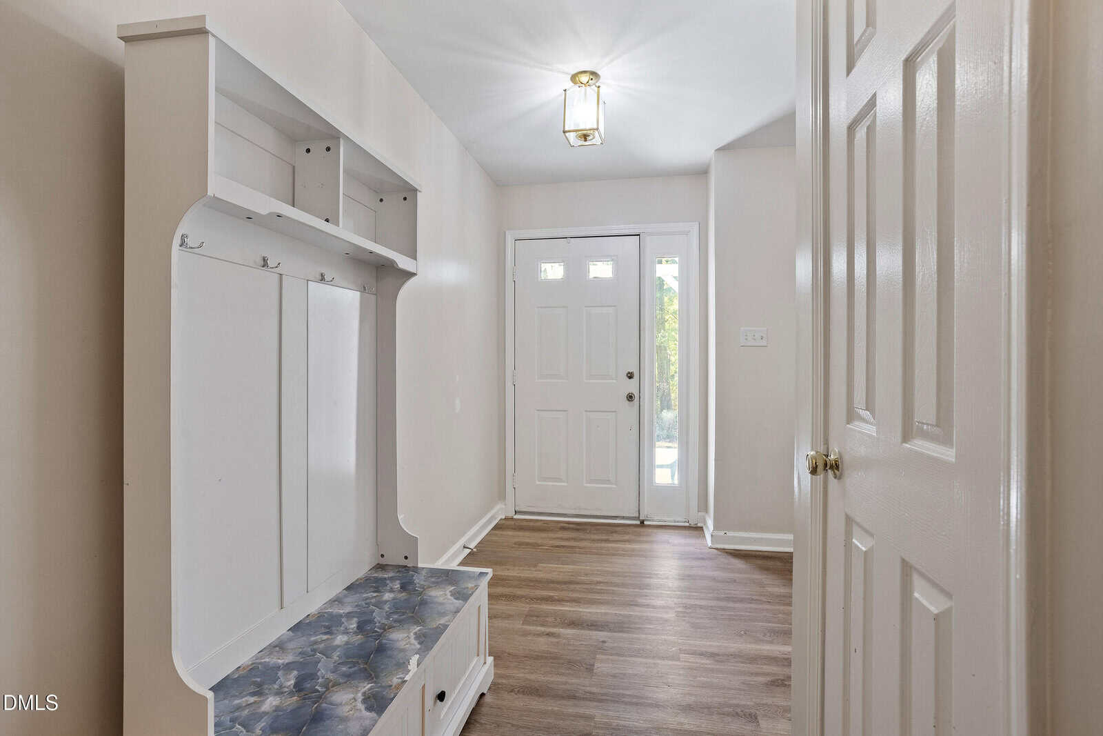 Bright entryway with white front door, side windows, light wood flooring, cream walls, and a built-in white hall tree with hooks, shelves, and a blue cushioned bench on the left side.