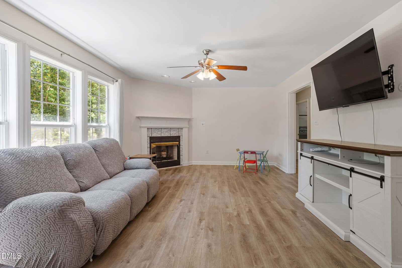 Bright living room with gray couch, ceiling fan, wall-mounted TV, fireplace, and kids’ table with chairs. Large windows let in natural light. Wood flooring throughout.