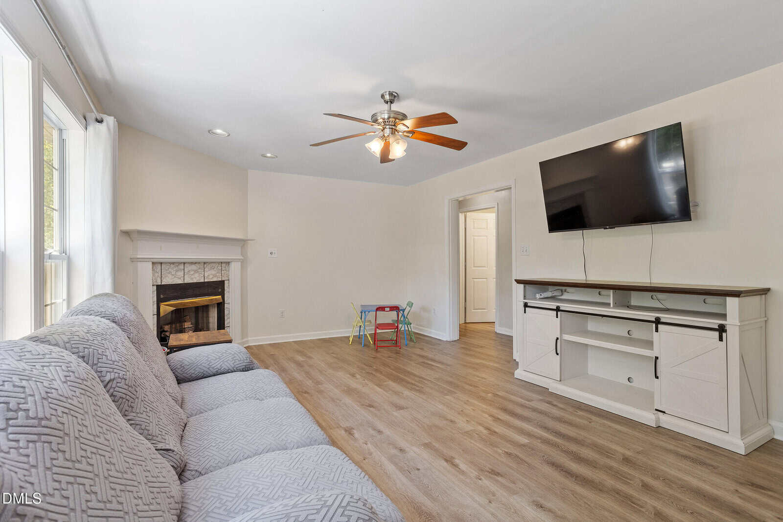 Bright living room with wood flooring, gray sofa, wall-mounted TV, white entertainment console, fireplace, and a small kids’ table with chairs near a window with light curtains. Ceiling fan with light in the center.