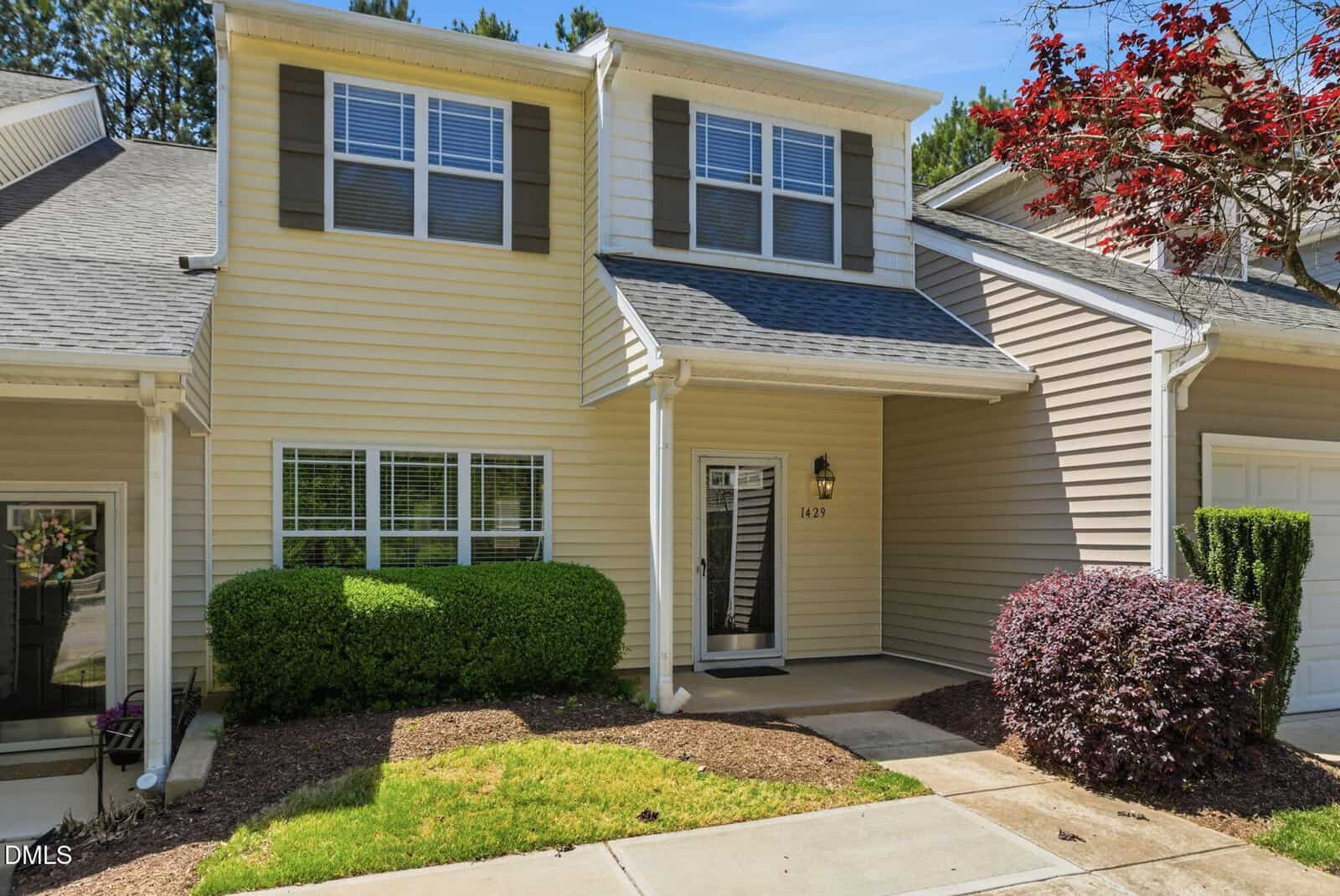 A beige two-story townhouse with white trim at 1429 Montonia Street, Wake Forest, features a covered entryway, green shrubs, a small tree, and a concrete pathway leading to the front door on a sunny day.