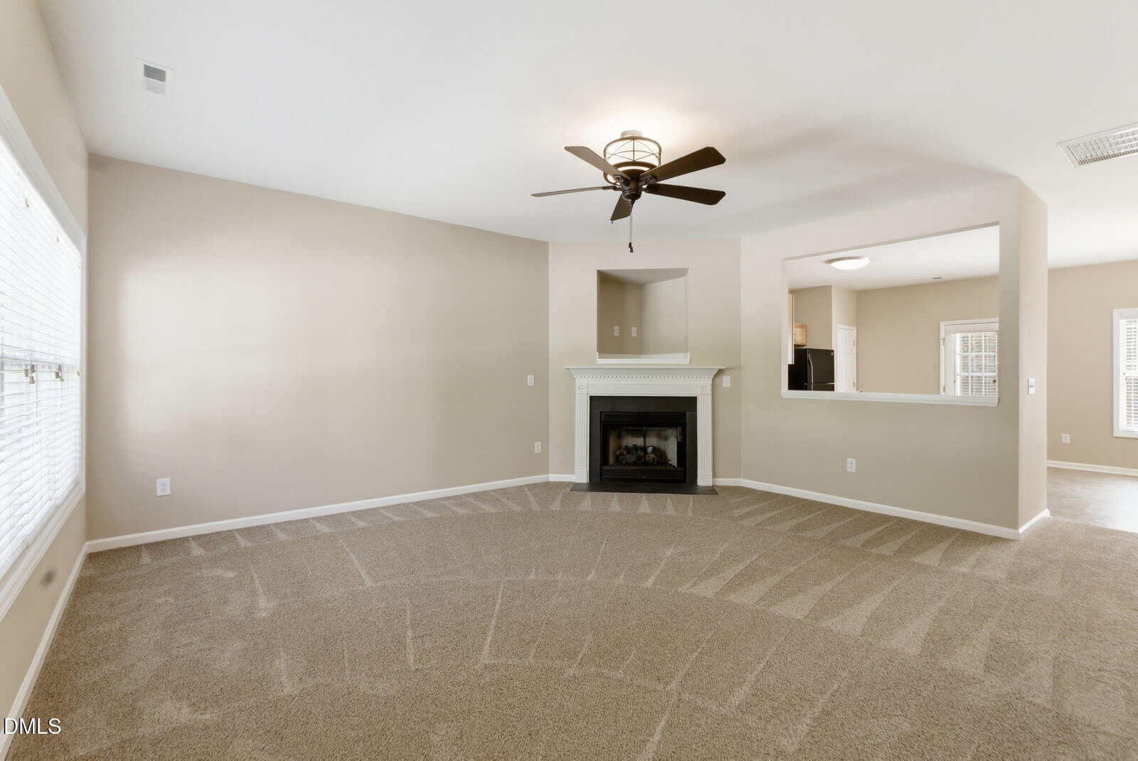 A spacious living room with beige walls, freshly vacuumed carpet, a white fireplace, ceiling fan, and large windows letting in natural light connects to a bright adjacent room through a wide open wall.