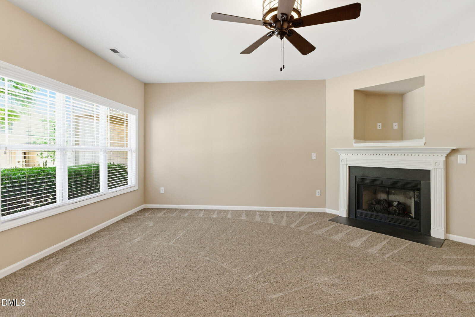 A spacious living room with beige walls, large windows with white blinds, a ceiling fan, a white fireplace, and freshly vacuumed carpet. Natural light enters from the left side.