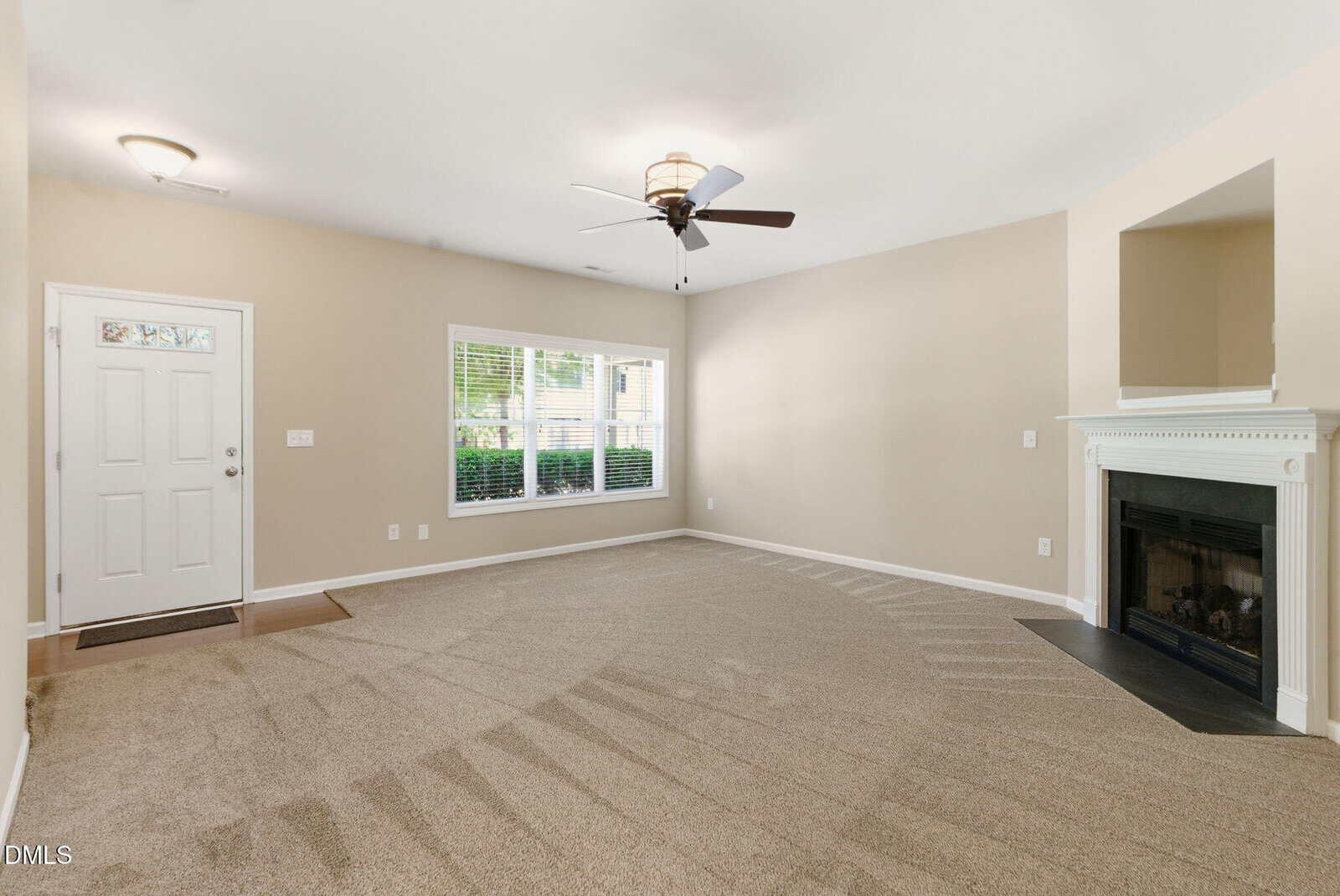 A spacious, empty living room with beige walls, clean carpet, a ceiling fan, large window, white front door, and a white fireplace with a black insert on the right wall.
