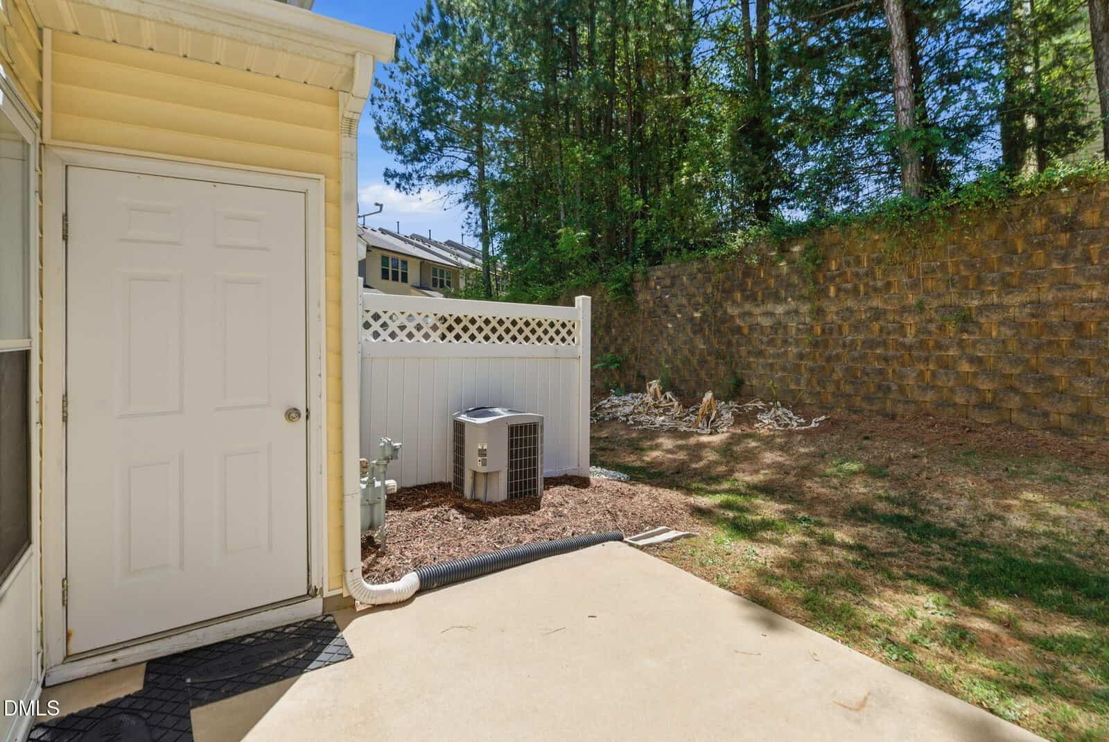 A small backyard with a concrete patio, a white door, an air conditioning unit, a white fence, and a grassy area bordered by a tall stone retaining wall and trees.