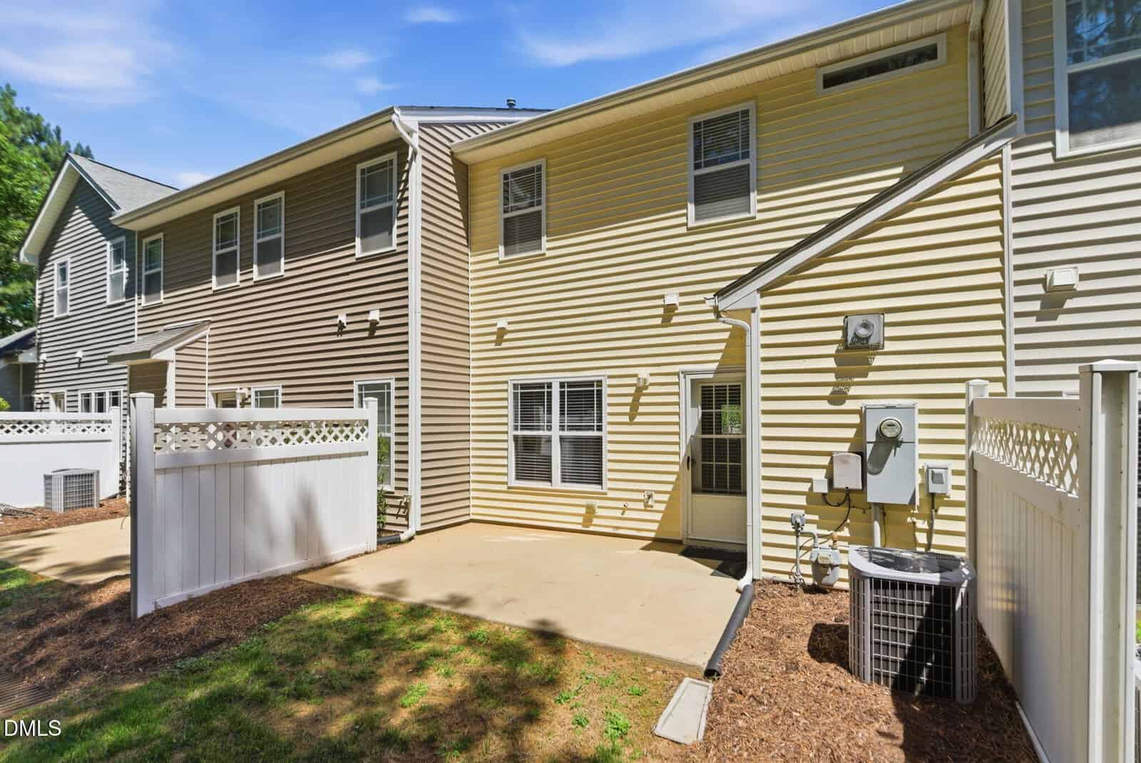 A two-story townhouse with beige siding, several windows, a concrete patio, a white fence, and an outdoor air conditioning unit on a sunny day.
