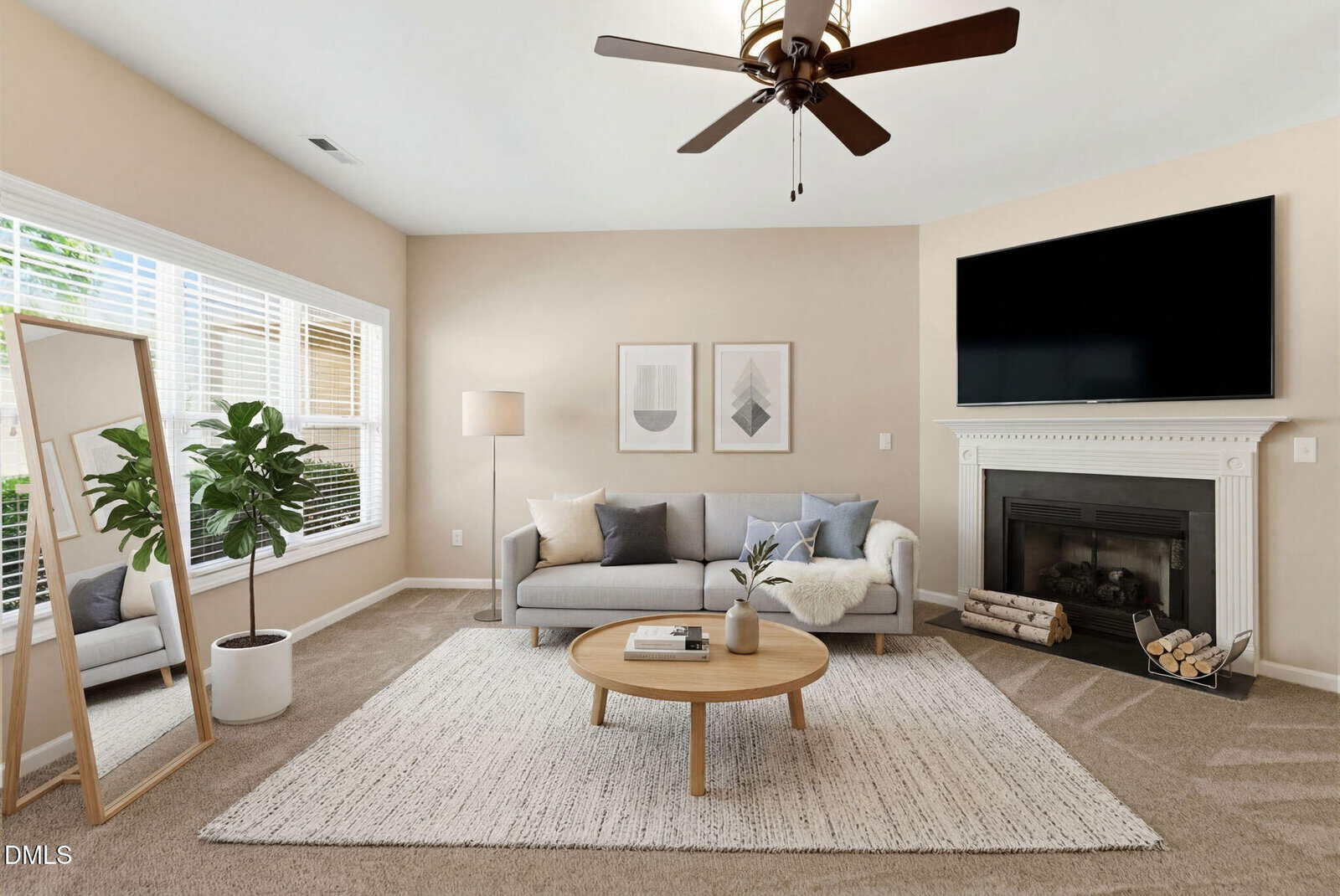 A modern living room with a gray sofa, round wooden coffee table, large rug, potted plant, standing mirror, and a fireplace with a TV above it. Large windows let in natural light. Neutral decor with minimalist artwork.