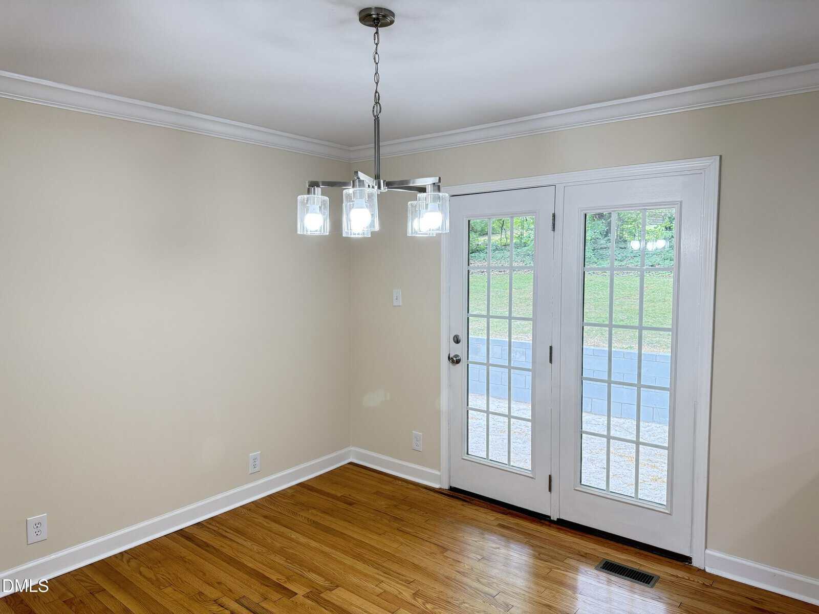 A bright, empty room with hardwood floors, cream-colored walls, a modern ceiling light fixture, and French doors leading outside to a patio with a grassy area and retaining wall.