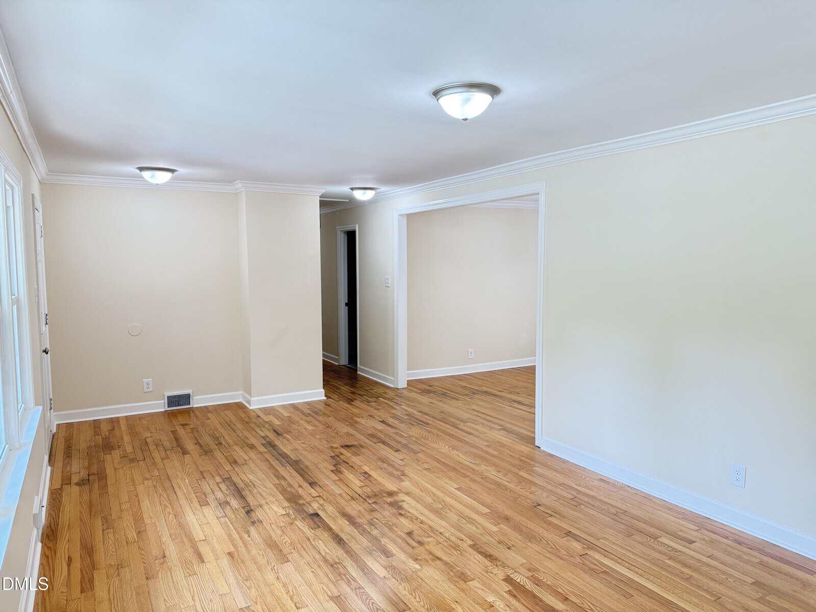 A bright, empty room with light beige walls, hardwood floors, white trim, and ceiling lights. There are large windows on the left and an open doorway leading to another room.