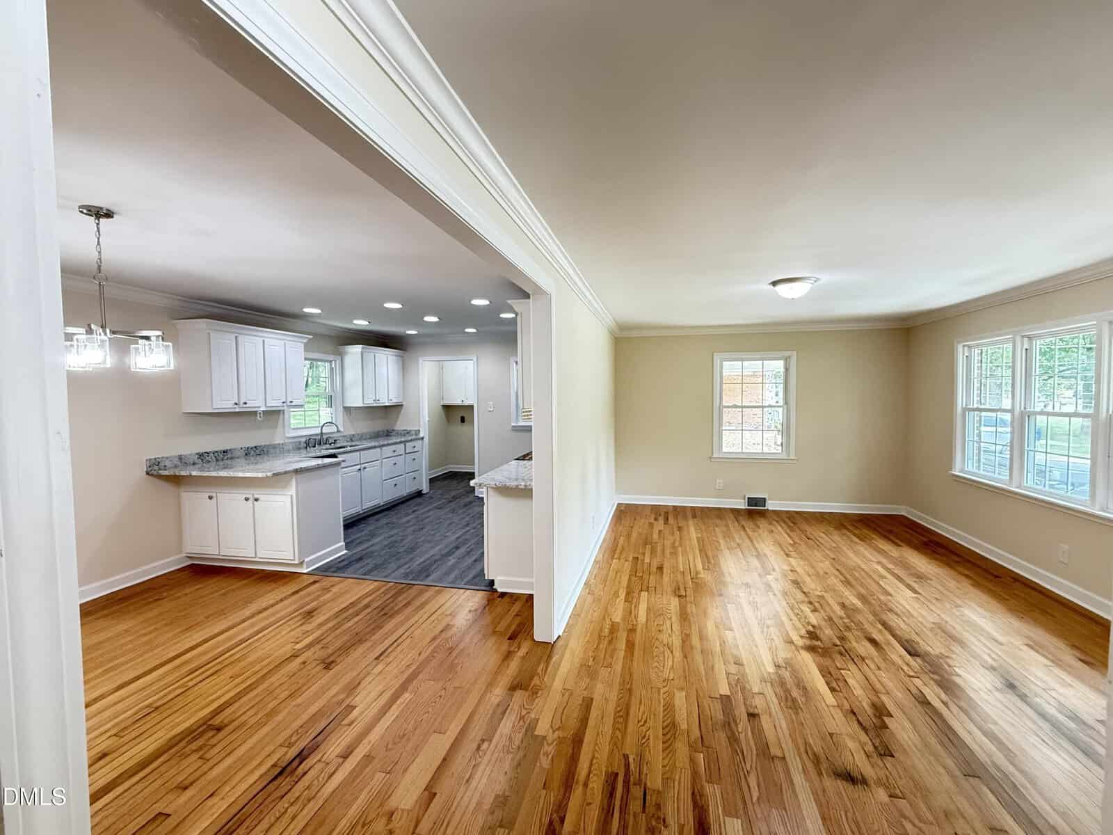 Open-concept interior featuring a bright kitchen with white cabinets and granite countertops, adjacent to a spacious living area with hardwood floors and large windows letting in natural light.