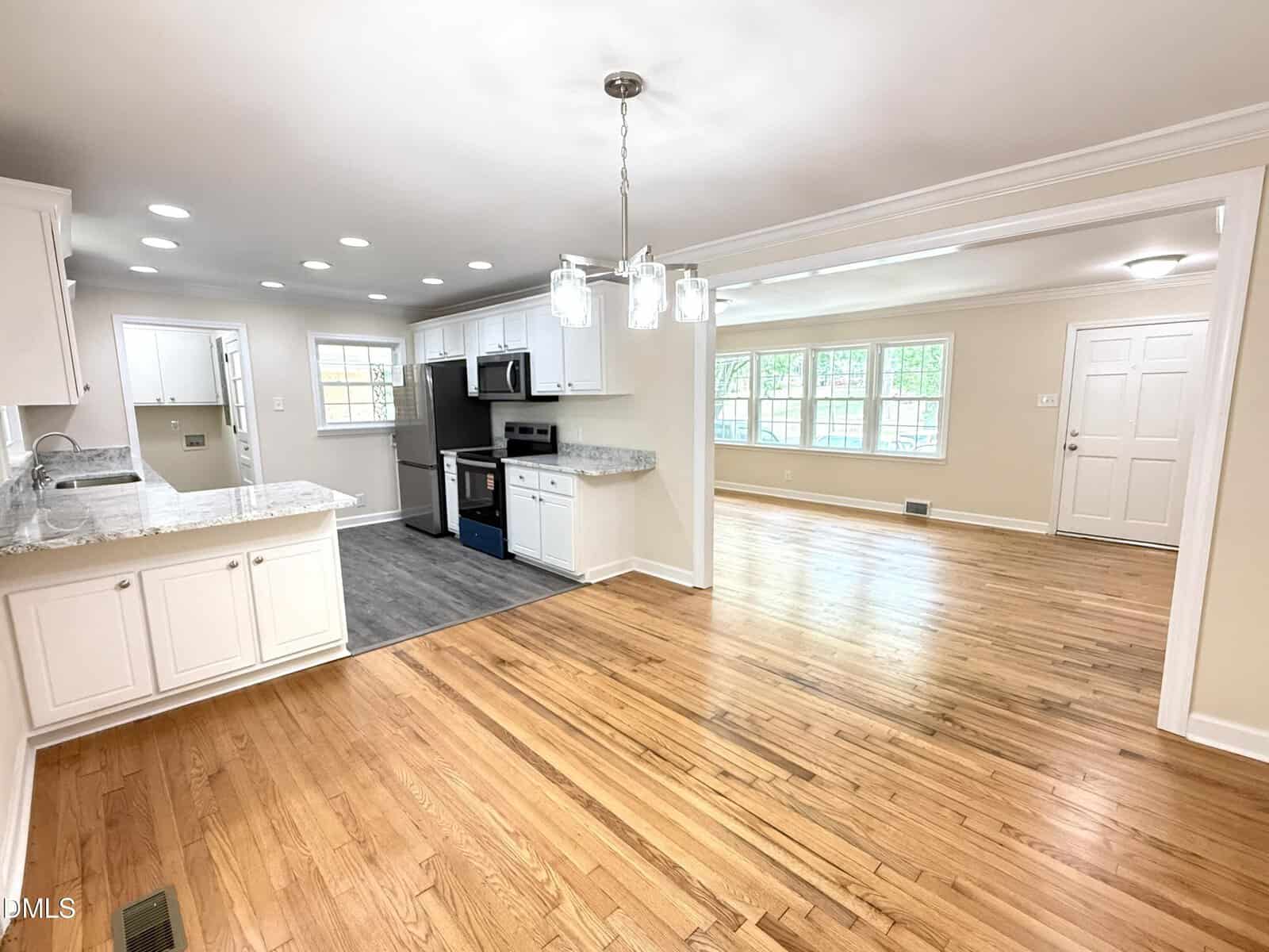 Bright, open kitchen and dining area with white cabinets, stainless steel appliances, marble countertops, wood floors, and large windows letting in natural light. Adjacent living room shown with matching hardwood floors.