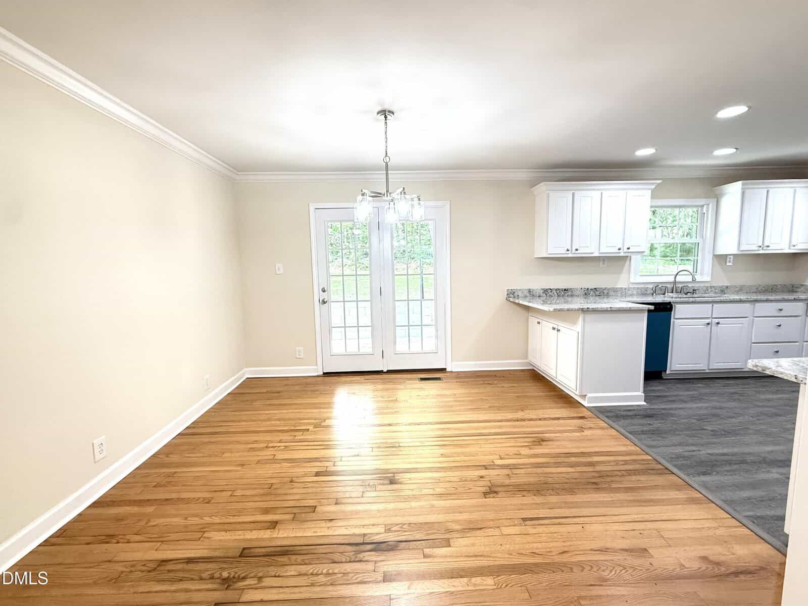 Bright dining area with hardwood floors, a chandelier, glass doors to the outside, and an adjacent white kitchen with granite countertops and gray flooring.