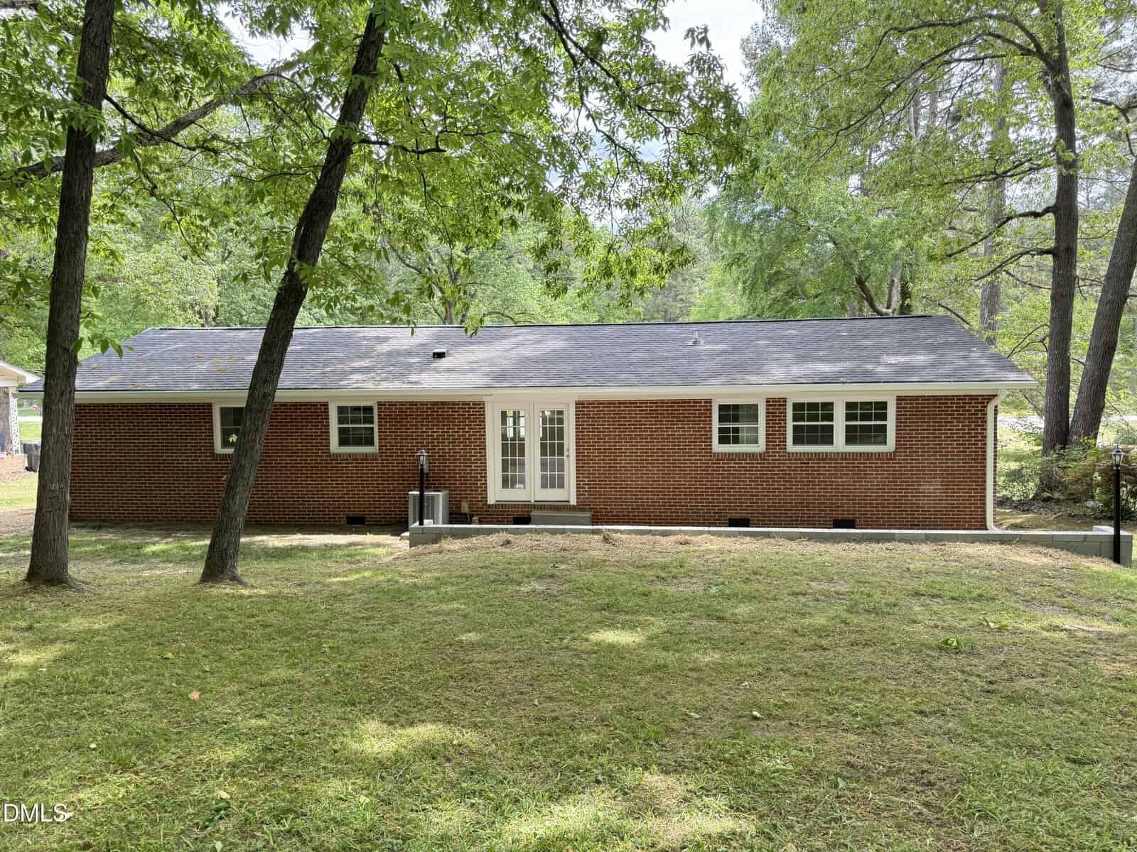 A single-story red brick house with a gray shingle roof and white-trimmed windows and doors, seen from the backyard. The yard is grassy with several trees and a small concrete step leading to the back door.