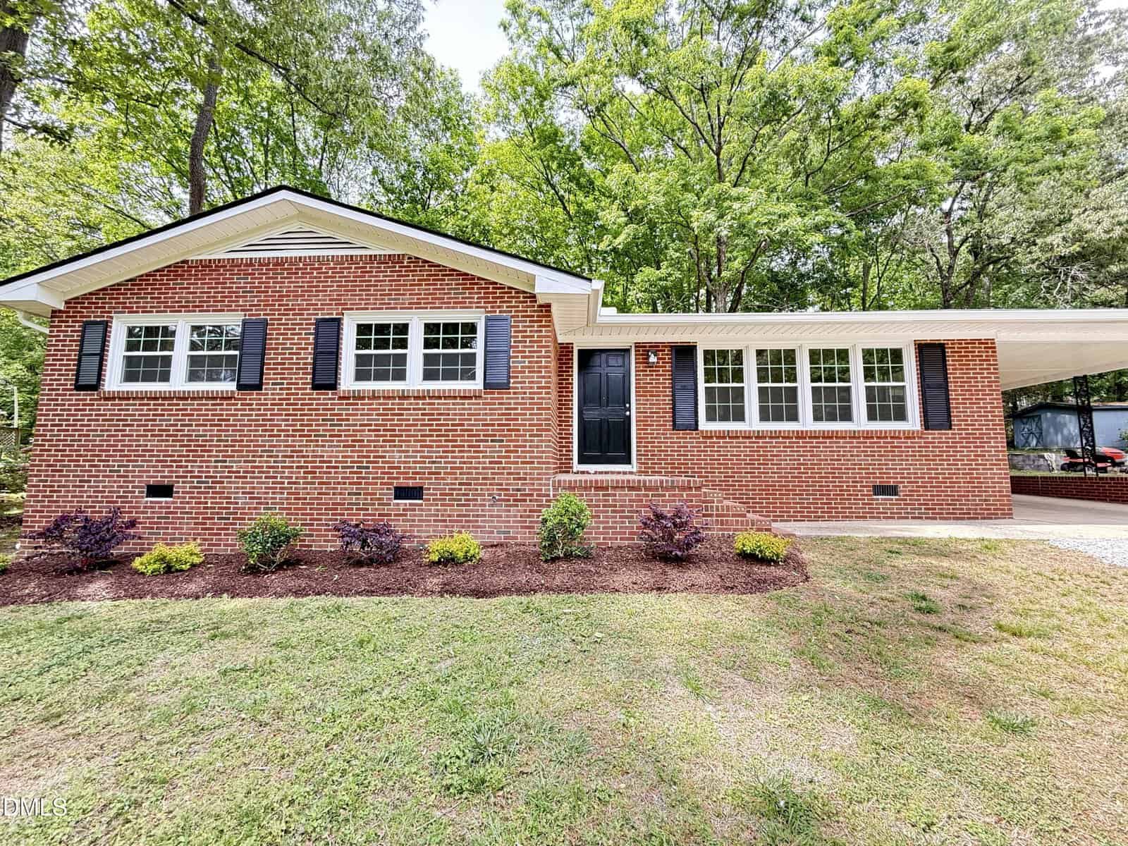 A single-story brick house with black shutters, white trim, and a covered carport. There are shrubs planted in front, and the house is surrounded by green trees and a grassy yard.