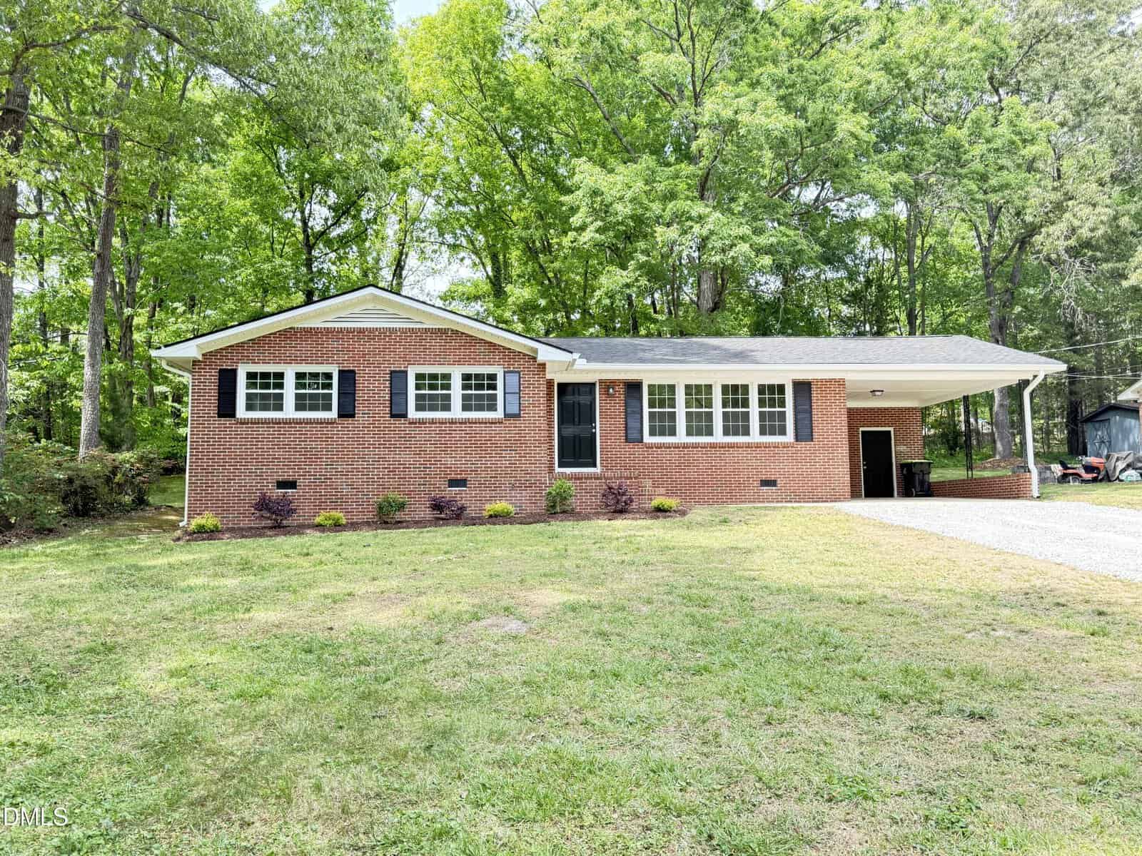 A single-story brick house with black shutters, large front windows, and a covered carport. The house is surrounded by green grass, shrubs, and tall trees in the background.