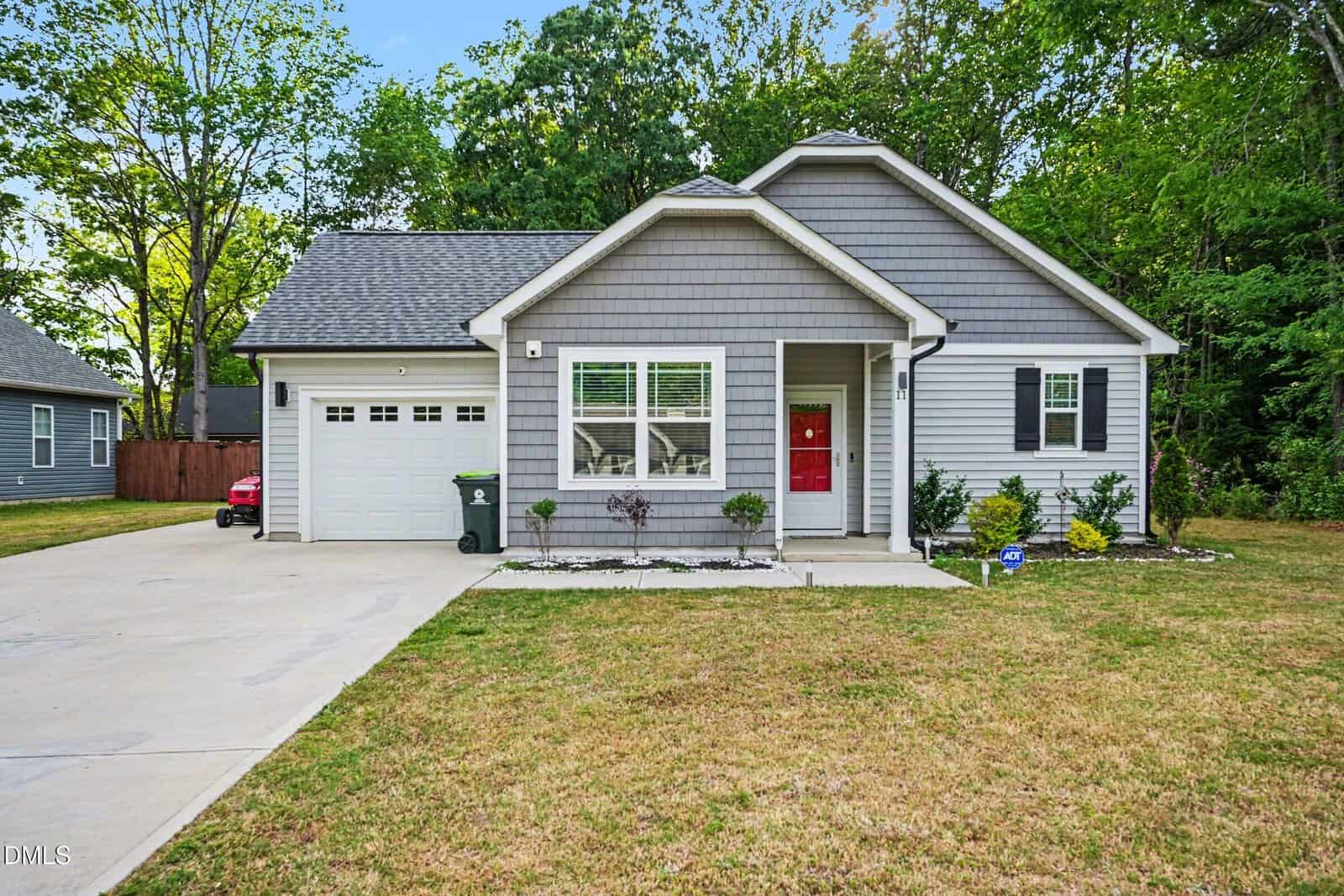 A single-story gray house with white trim and a red front door, attached one-car garage, driveway, small front garden, and a grassy yard bordered by trees.
