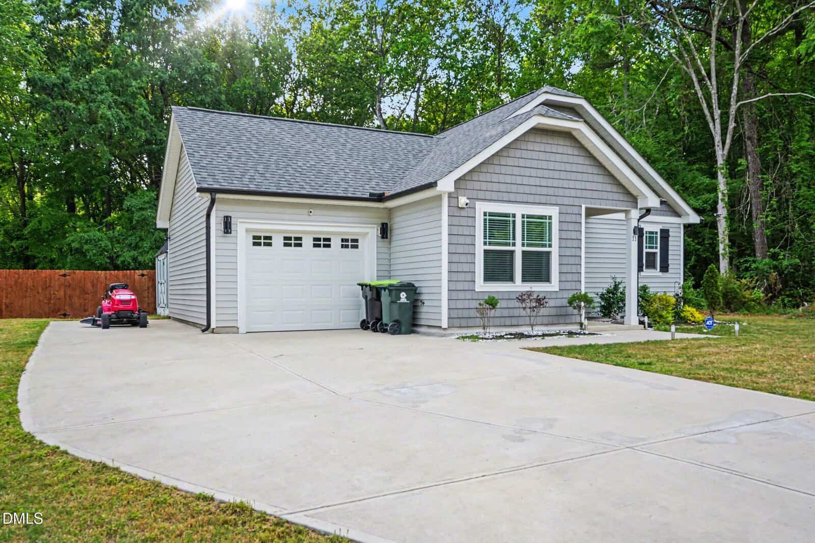 A small, modern gray house with white trim and a single-car garage sits beside a wide driveway. A toy car and trash bins are near the garage; trees and a wooden fence border the backyard.