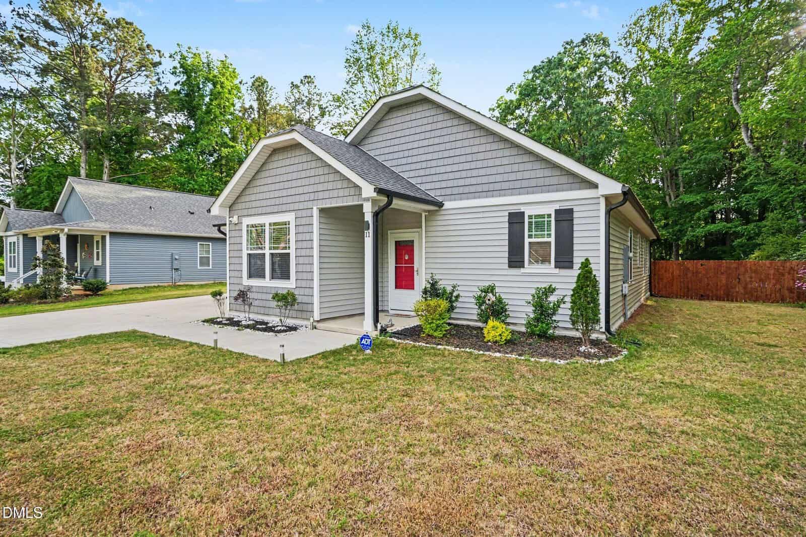 Single-story gray house with a red front door, small front garden, and concrete driveway. The home has white trim, a manicured lawn, and is surrounded by trees and a wooden fence.