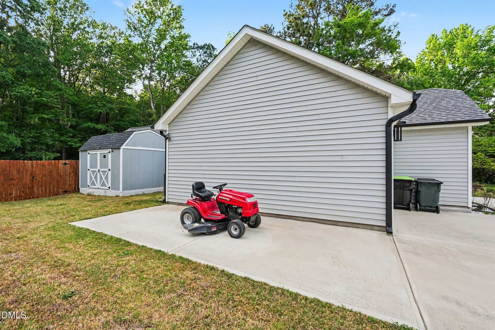 A red riding lawn mower sits on a concrete patio next to a light gray house with white trim. A small gray storage shed and wooden fence are visible in the background. Green trees surround the yard.