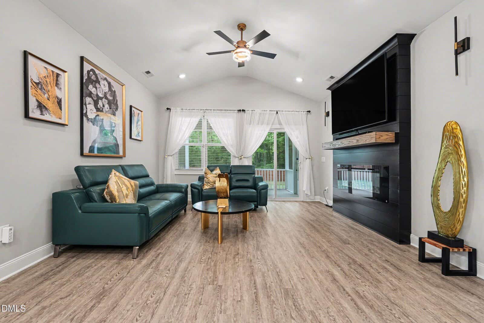 Modern living room with a dark teal leather sofa and love seat facing a black fireplace with a mounted TV. A gold abstract sculpture sits on a bench beside the fireplace.