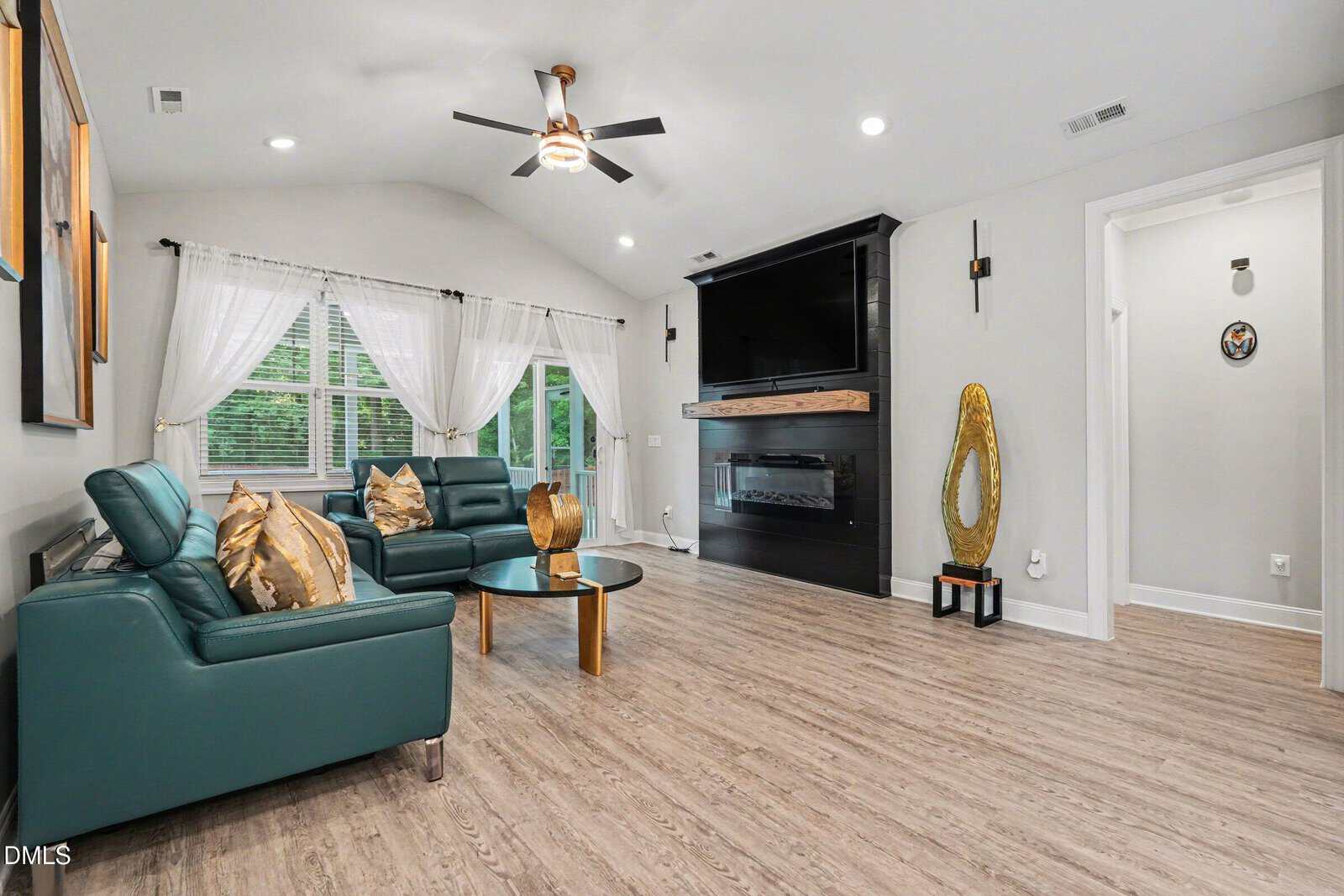Modern living room with teal sofas, glass coffee table, large windows with white curtains, wood floor, wall-mounted TV above a fireplace, and decorative gold sculptures. Hallway visible to the right.